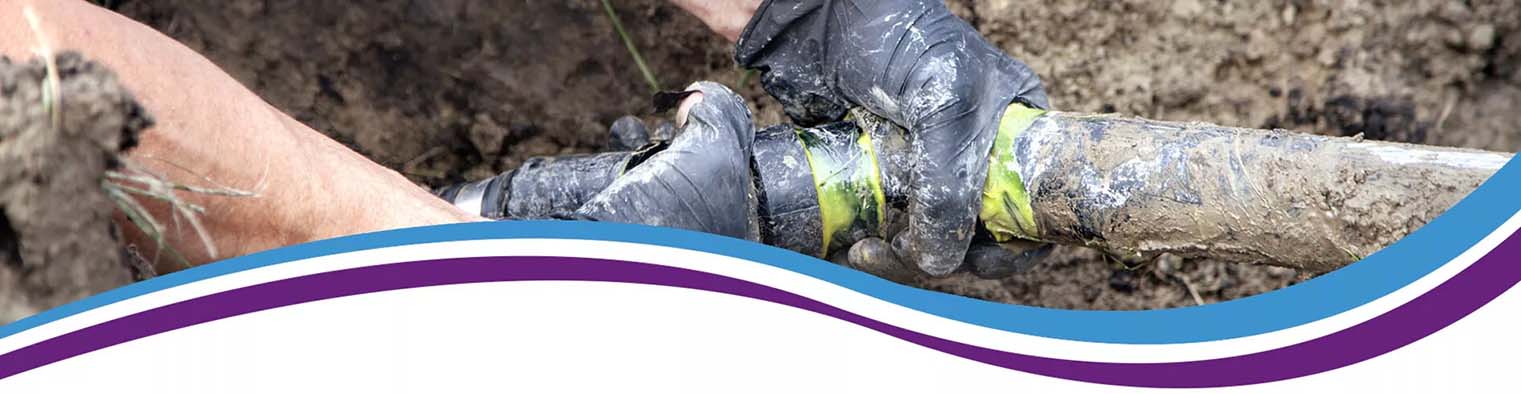 Image shows gloved hands working on a muddy pipe in a trench