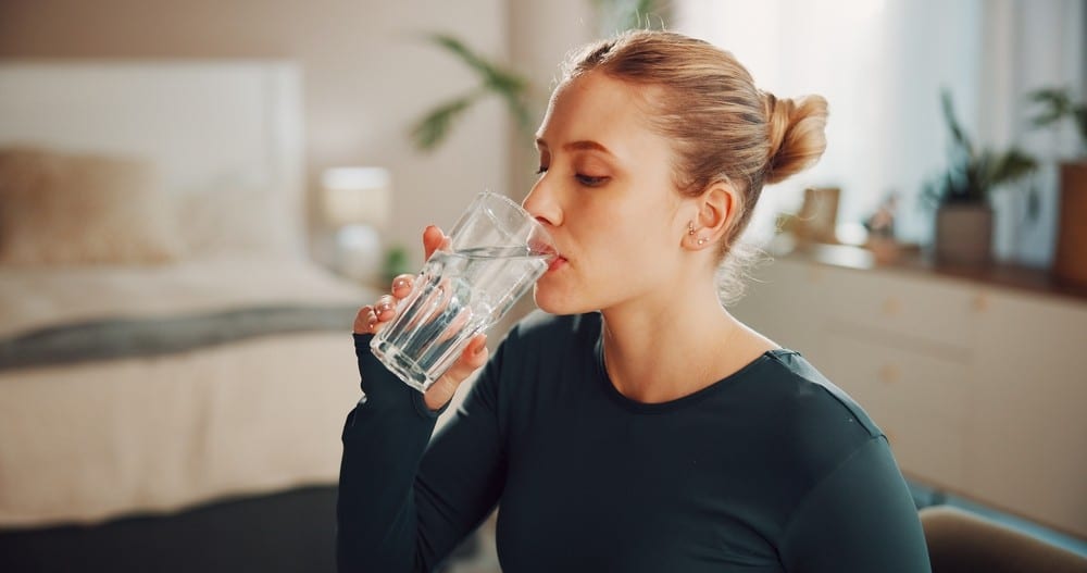 A woman drinking water from a glass, while in her kitchen.